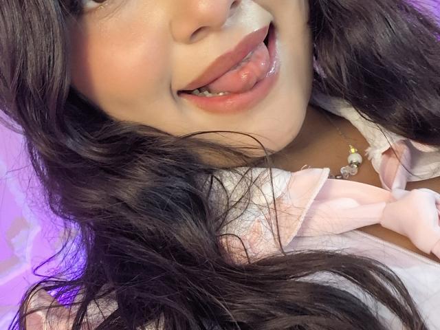 NekoPam with long black hair, light skin, and pink lips, tongue out, showing teeth, wearing a pink top with a bow, necklace, and a cute, playful expression.