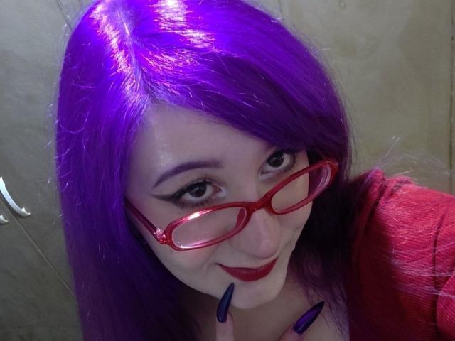 DulcieStuckert with purple hair, red glasses, and red lipstick, smiling at the camera, finger touching her lips, wearing a red top, with purple nails.