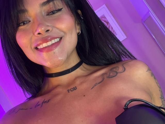 MajoJones, con cabello negro, sonriendo, mostrando sus pechos, con tatuajes en el pecho, un collar, un piercing en la nariz y una pequeña cantidad de semen en la cara. Luz púrpura en el fondo..