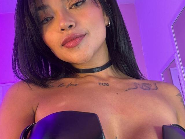 MajoJones with black hair, choker, and tattoos, showing bare tits with black straps, pink and purple lighting, and a confident smile.