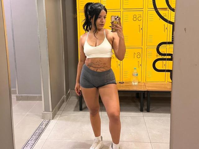 Sophie Soler, met zwart haar in een staart, draagt een witte sportbeha en grijze shorts, waarbij haar getoonde buikspieren en witte sneakers zichtbaar zijn. Ze maakt een spiegelzelfportret in een sportschool..