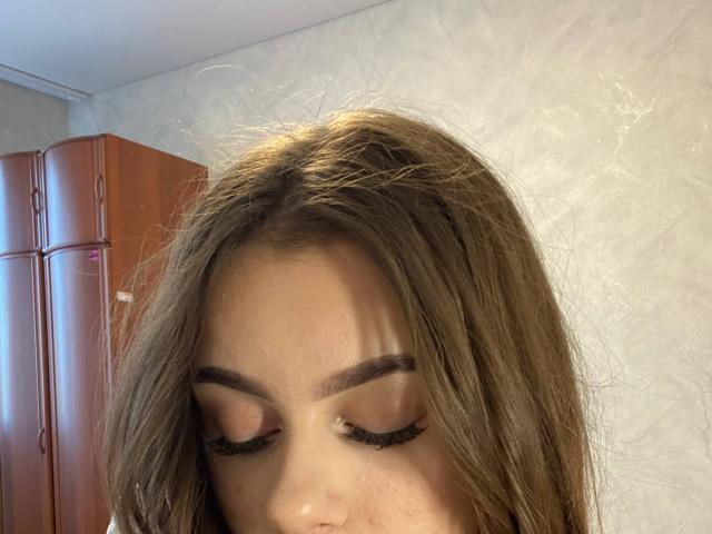 KimberlyZampieri's face, brown hair, closed eyes, long eyelashes, and light brown eyebrows, with a wooden fridge on the left and a textured white wall in the background.