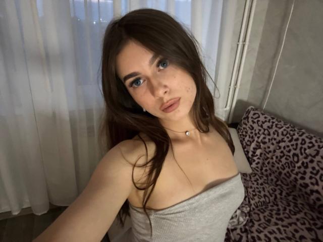 KimberlyZampieri with long brown hair, blue eyes, and a gray strapless top, showing her small tits and a neutral expression, standing near a leopard-print pillow.