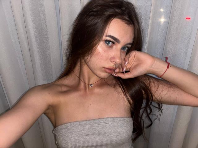 KimberlyZampieri with long brown hair, blue eyes, and freckles, wearing a gray strapless top, touching her face with a red bracelet on her wrist.