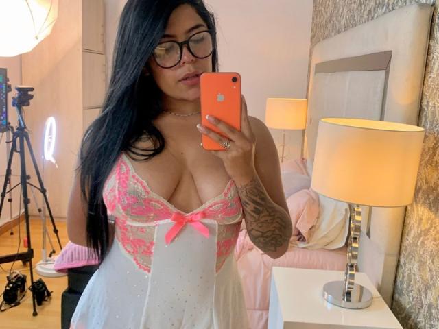 CatalinaReiyna with glasses, black hair, and tattoos, wearing a pink lace and white babydoll with a pink bow, taking a selfie in a bedroom with a lamp and bed.
