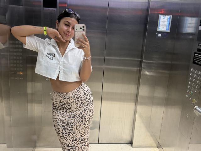 SarahPrincess, avec ses cheveux foncés, une chemise blanche courte qui dévoile sa poitrine, et un pantalon à motifs léopard, prend un selfie dans un ascenseur en métal. Elle sourit et porte des lunettes de soleil sur la tête..