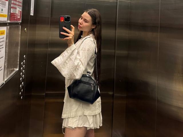 Azzie Stelman, con lunghi capelli castani, indossa un abito bianco ampio e una borsa nera, e sta scattando un selfie allo specchio in un ascensore metallico, tenendo in mano un telefono con una custodia rossa..