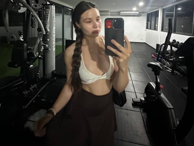 AzzieStelman maakt een selfie voor een spiegel in de sportschool, ze draagt een witte beha die haar grote borsten laat zien en een bruine rok, met lange vlechten, ze houdt een telefoon vast en heeft een lichtjes fronsende uitdrukking..
