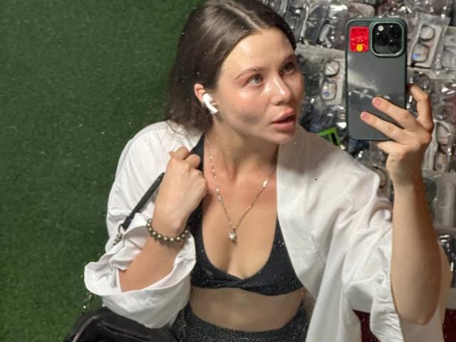 Azzie Stelman, con un seno di media grandezza, indossa un top da bikini nero, una camicia bianca e auricolari, e sta scattando un selfie allo specchio, tenendo in mano un telefono con una custodia verde..
