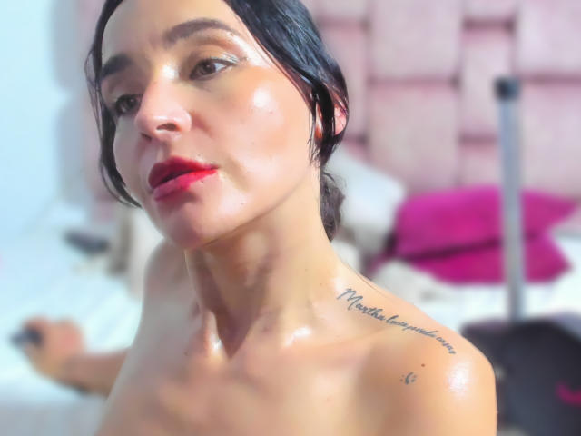 MindyLove has shiny skin, black hair, and red lipstick, with a tattoo on her neck. She's topless, showing her boobs, with a slight shine on her chest.