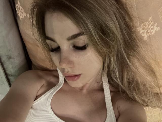 MayaCatty with closed eyes, wearing a white tank top, showing her tits, with light brown hair, lying down, soft pink pillow, subtle makeup, and slightly parted pink lips.