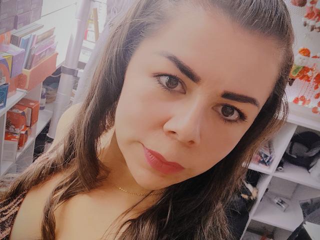 EmiliaMilf with brown eyes, medium brown hair, and pink lips, wearing a gold necklace, has a cute, slightly smug expression, and is showing her tits in a selfie.