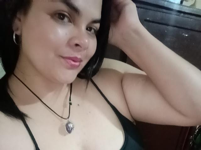 BiancaBeck tem a pele clara, cabelos pretos e está vestindo uma blusa verde com um colar. Ela está mostrando os seios e tem um sorriso fofo e delicado..