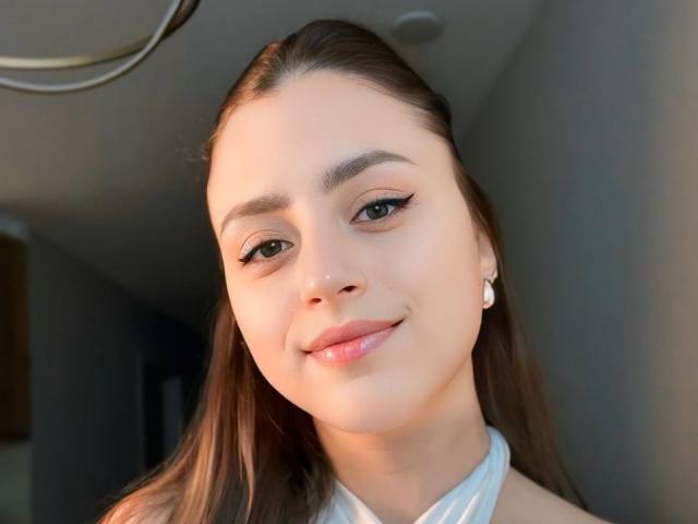 CarynHowkins tem uma pele lisa e clara, olhos castanhos e um leve sorriso. Ela tem cabelos castanhos longos, delineador, lábios rosados e uma blusa branca de alças. Ela está usando pequenos brincos de pérola..