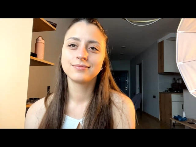 CarynHowkins heeft lang bruin haar, een subtiele glimlach en een schattig gezicht met een lichte huid, en draagt een witte top. Ze staat in een helder verlichte kamer met een ringlamp..