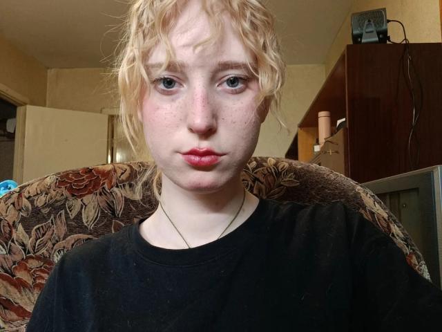 JennifferBreaz with curly blonde hair, pale skin, freckles, and red lips, wearing a black shirt, looking at the camera, and a thin necklace, in a cozy room.