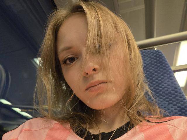 ShawandaClester has messy blonde hair covering one eye, a small nose, and is wearing a pink and black shirt, looking slightly bored with a subtle pout.