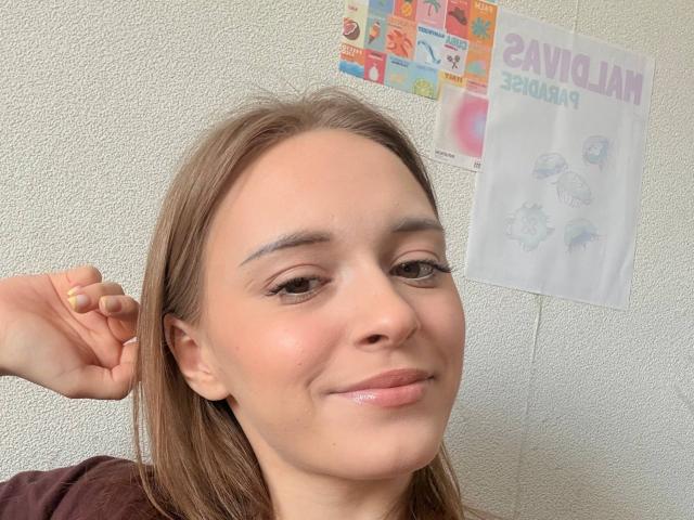 ShawandaClester heeft lichtbruin haar, bruine ogen en een lichte glimlach met glanzende lippen, ze houdt haar haar vast met een gele nagel en draagt een bruin shirt..