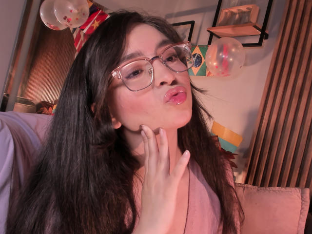 EvaMcAllister with long dark hair, wearing glasses, pouting lips, pink blush, and a pink top, touching her chin, surrounded by balloons and a party hat.