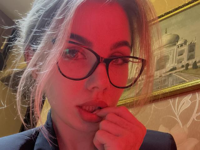 MayaCatty with red-tinted face, black glasses, and slightly parted lips, biting her finger. She has blonde hair in a messy ponytail, wearing a dark top.