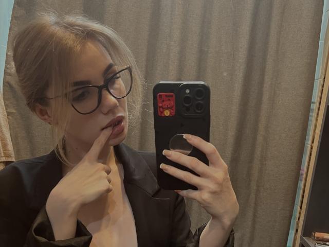MayaCatty, avec des cheveux blonds attachés en un chignon désordonné, portant des lunettes et un blazer noir, mordant sa lèvre en prenant un selfie devant un miroir avec un téléphone noir..