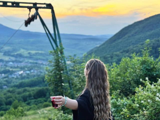 Niferttiti, com cabelos castanhos ondulados, vestindo uma blusa preta, segurando um café vermelho, com vista para montanhas, uma floresta verde e o pôr do sol. Um teleférico está suspenso ao fundo. As nádegas de Niferttiti estão levemente visíveis..