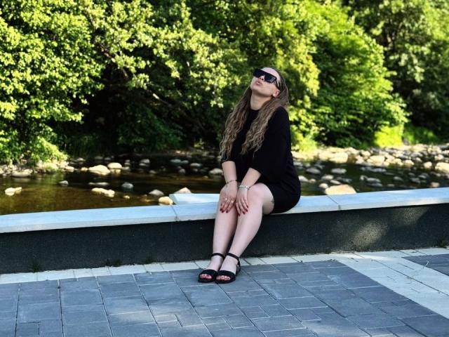Niferttiti está sentada en un banco negro, vestida con un vestido negro, sandalias negras y gafas de sol oscuras, con el pelo largo y ondulado de color marrón y uñas rojas, en un parque soleado..