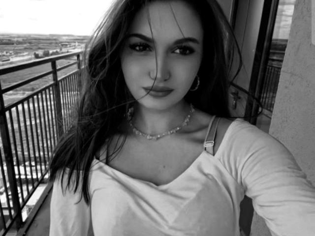 RamonaOsazuwa tem cabelo escuro, usa um piercing no nariz e veste uma camisa branca de manga comprida com um colar, mostrando levemente os seios. Selfie em preto e branco, olhando diretamente para a câmera..