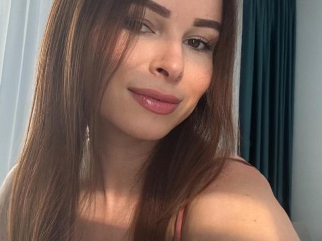 AnnaBlossom a les cheveux bruns, les lèvres roses et un sourire charmant. On aperçoit légèrement ses seins sous un haut rouge, et elle regarde l'appareil photo. Elle a un look sexy et naturel..