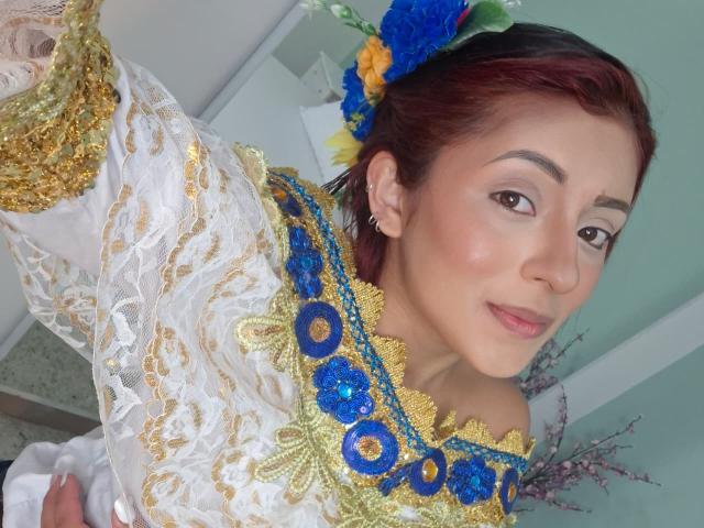 LizBonnet with red-brown hair, wearing a white and gold lace dress with blue and gold floral embroidery, holding a selfie stick, blue and yellow flower in her hair.