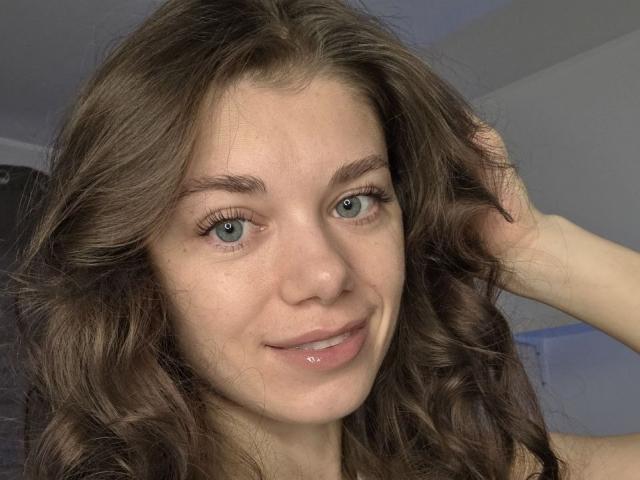 YourRedFlag tem cabelos castanhos ondulados, olhos azuis e um leve sorriso com lábios brilhantes. Sua mão direita está no cabelo, mostrando uma expressão fofa e relaxada..