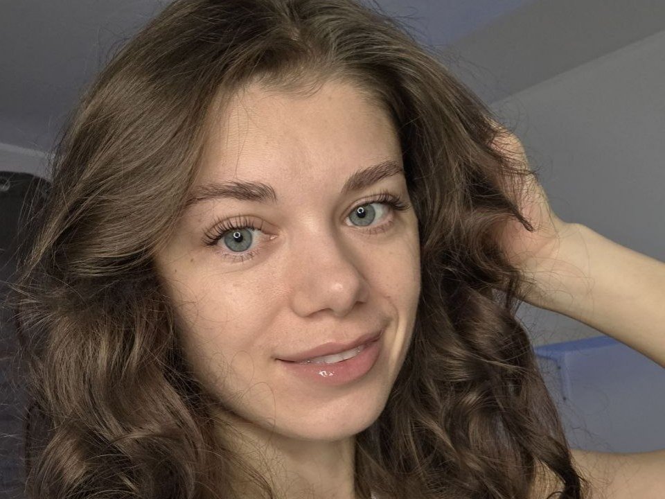 YourRedFlag hat welliges, braunes Haar, blaue Augen und ein leichtes Lächeln mit glänzlichen Lippen. Ihre rechte Hand ist in ihrem Haar, was einen süßen, entspannten Ausdruck zeigt..