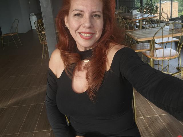 Khharha with red hair, smiling, wearing a black off-shoulder top, showing her boobs, in a sunlit cafe, taking a selfie, her right arm raised.
