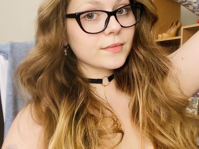 KarKutie, met golvend bruin haar, zwarte bril en een zwarte choker, heeft een neutrale uitdrukking en zichtbare tatoeages op haar arm. Haar borsten zijn gedeeltelijk zichtbaar..