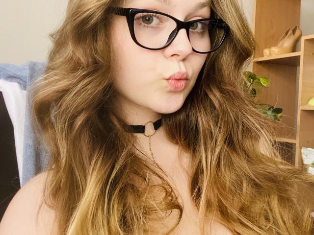 KarKutie met lang, golvend blond haar, zwarte bril en een zwarte choker, die haar roze lippen opzettelijk naar voren duwt, waarbij haar blote schouders en een ketting zichtbaar zijn..