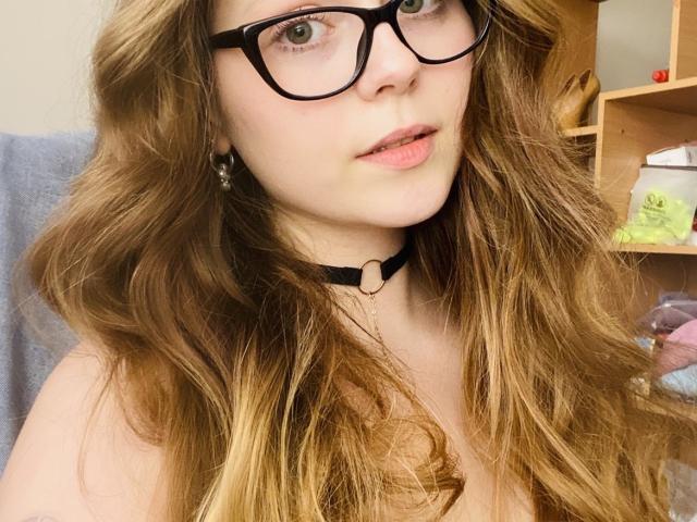 KarKutie met lang, golvend bruin haar, zwarte bril en een zwarte choker, kijkt iets omhoog met roze lippen, waarbij haar borsten zichtbaar zijn. Ze heeft een schattelijk, sexy gezicht..