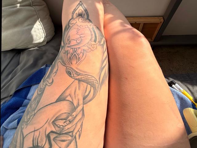 La gamba tatuata di Prettyjosi-hot, con dettagliate linee che raffigurano un serpente e un motivo ornamentale, la luce del sole che proietta ombre, la gamba scoperta accanto, un asciugamano blu sullo sfondo..