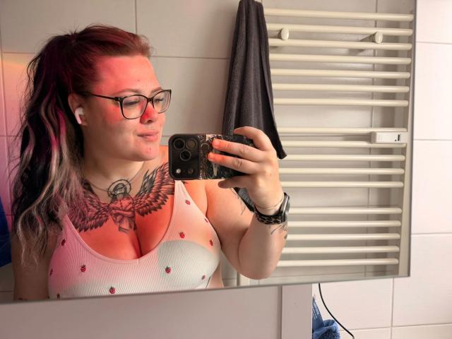 Prettyjosi-hot with glasses, white tank top showing big tits, red tattoo on chest, holding phone, red light reflection, black hair in ponytail, white earbuds, white towel rack.