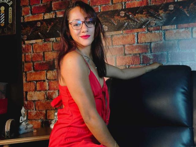 SamanthaCorine, avec de longs cheveux bruns, des lunettes, une robe rouge et du rouge à lèvres, assise sur une chaise en cuir noir, le bras appuyé en arrière, souriante, montrant ses seins, avec un mur de briques en arrière-plan..