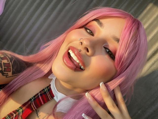 ValkaGreen, com cabelo rosa, língua de fora, mostrando um piercing de lábio prateado e um piercing no nariz, vestindo uma camisa xadrez, gola branca, e segurando o cabelo com os dedos..