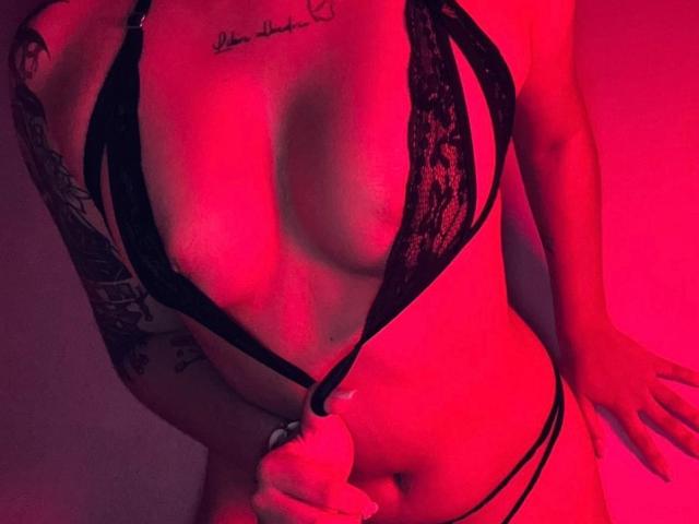 ValkaGreen vestindo um conjunto de lingerie preta com detalhes em renda, iluminação vermelha, com tatuagens visíveis, segurando um fio preto perto da vulva, com os seios e a mão na imagem..