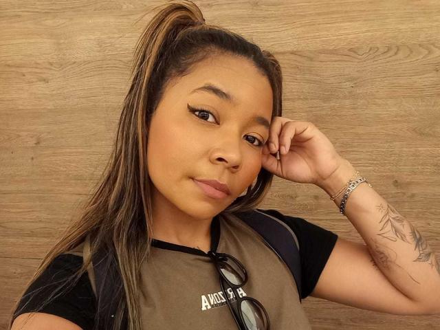 XinemaBonet con la pelle marrone, una lunga coda di cavallo marrone, eyeliner nero, indossa una camicia nera e marrone, tiene degli occhiali da sole, ha un tatuaggio sul braccio destro, sfondo in legno, trucco leggero, guarda la fotocamera..