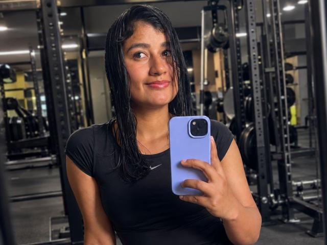 Adara Rivat, con el cabello negro y mojado, vestida con una camiseta negra ajustada de Nike, sosteniendo un teléfono azul, sonriendo en un gimnasio, mostrando unos glúteos lindos y curvilíneos en el reflejo del espejo..
