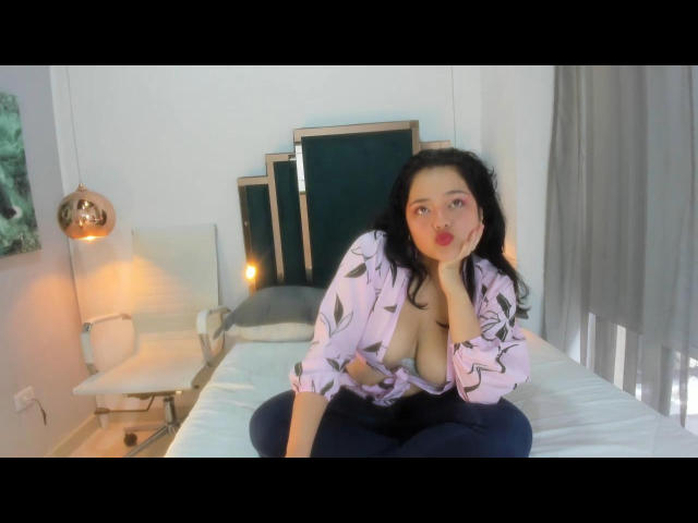 Esme Miller, con cabello negro y rizado, una camisa rosa con estampado floral y pantalones azules, sentada en una cama, tocándose los labios cubiertos de lápiz labial rojo, mostrando sus grandes senos, mirando a la cámara..