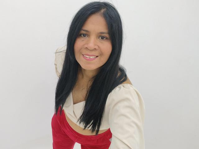Rhouss, avec ses longs cheveux noirs, souriante, portant un haut crème à une épaule et un pantalon rouge en velours, penchée en avant, montrant son joli visage et sa petite poitrine naturelle..