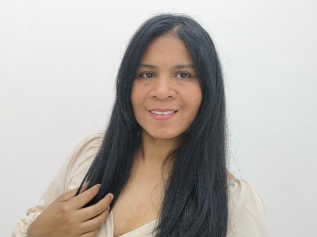 Rhouss a de longs cheveux noirs, une peau brun clair, de petites seins et un sourire charmant. Elle porte une chemise beige et tient sa chemise d'une main..