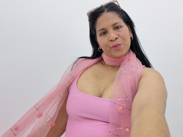 Rhouss, eine kurvige lateinamerikanische Frau mit langen schwarzen Haaren, pinkem Lippenstift und großen Ohrringen, trägt ein pinkes Tanktop und einen durchsichtigen, pinken Schal mit floralen Mustern, der ihre Brüste zeigt..
