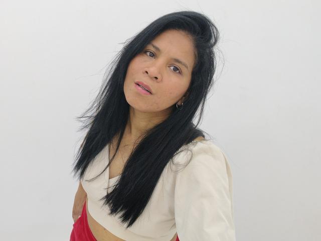 Rhouss a de longs cheveux noirs, des yeux marrons et une peau claire. Elle porte un haut blanc avec un tissu rouge, et ses petites seins sont légèrement visibles. Elle regarde l'appareil photo..