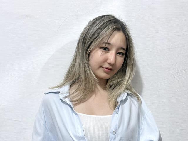 Amiee Shibata, con cabello rubio, vestida con una camisa blanca de botones abierta sobre un top blanco, tiene una sonrisa encantadora y un maquillaje sutil, y está de pie frente a una pared blanca lisa..