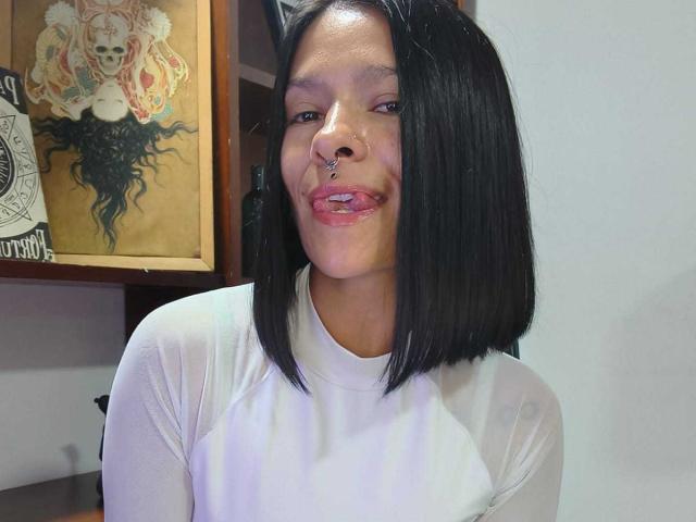 AmelieBlake with black bob, white shirt, nose ring, and tongue out, sitting, showing her tits slightly through sheer shirt. Tattooed wall art in background.